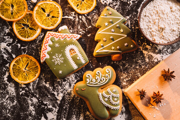 Festive decorated bakery. Christmas gingerbread cookies. Homemade culinary. Figured icing pastries house deer holiday tree flour powder dried orange spices on black desk.