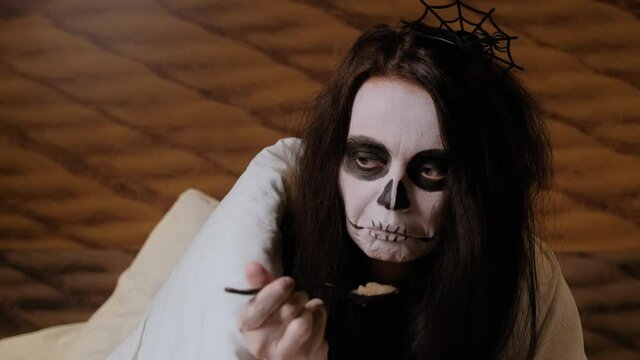 A girl in the form of a cute zombie sits on the bed and at first hesitantly, and then quickly and with pleasure, she eats the brain jam from the jar with a spoon. Halloween Eve.Festive humor. Close-up