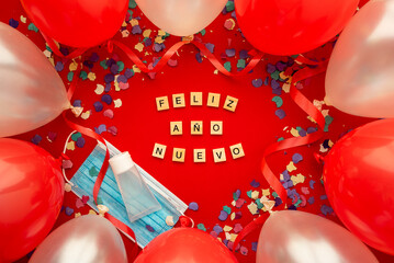 Party background with Coronavirus and happy new year written in Spanish with wooden letters and decorated with face mask, hydroalcoholic gel, colored balloons and confeti. Covid and party concept.