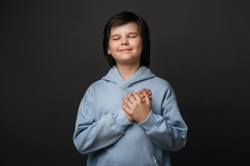 Cheerful boy 10-12 years old smiles gently and presses palms to heart. Positive emotions. Studio shot, gray background