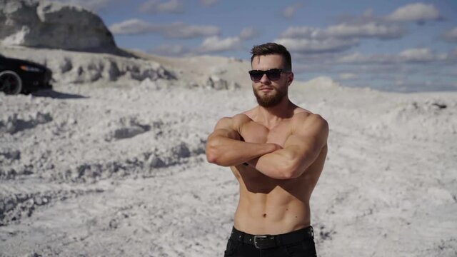 Portrait of shirtless athlete. Handsome guy with beard in sunglasses with muscular body posing on white canyon in summer. Camera motion around.