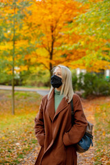 Style blonde in face mask and coat on autumn park with maples on background