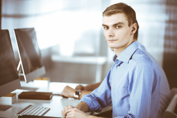 Two young confident businessmen, sitting at the desk in a modern office, while working on the...