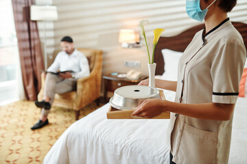 Maid in protective mask bringing hot breakfast and fresh flowers in hotel room of guest