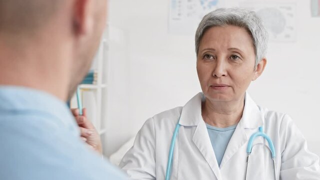Medium over-the-shoulder close-up of grey-haired Asian female neurologist sitting in medical office making unrecognizable patient watch pen moving for oculomotor system checking
