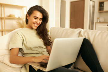Adorable Arabic girl in casual clothes relaxing on couch using laptop, looking at screen with smile, chatting with friends distantly, via group video conference call, enjoying online communication