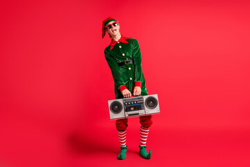 Full length photo cool elf hold boom box x-mas christmas party wear green costume isolated on red shine color background © deagreez
