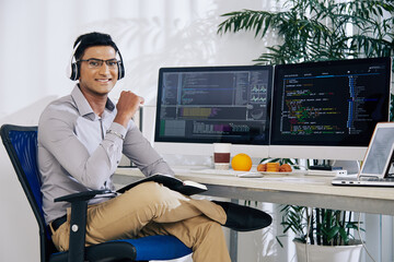 Portrait of smiling confident Indian coder sitting at his office desk with programming code on...
