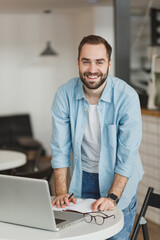 Cheerful attractive young man standing alone near table in coffee shop cafe restaurant indoors working or studying on laptop pc computer writing in notebook. Freelance mobile office business concept.