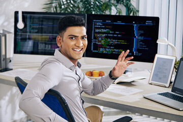 Happy excited Indian software developer sitting at office desk, turning back and waving at camera