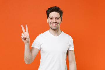 Smiling cheerful attractive young man 20s wearing basic casual white blank empty t-shirt standing showing victory sign looking camera isolated on bright orange colour wall background, studio portrait.