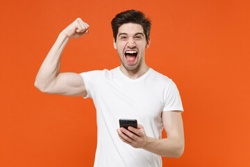 Overjoyed screaming young man in basic casual blank white t-shirt standing using mobile cell phone typing sms message doing winner gesture isolated on bright orange colour background studio portrait.