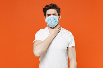 Sick displeased young man wearing white t-shirt sterile face mask to safe from coronavirus virus covid-19 during pandemic quarantine put hand on throat isolated on orange background studio portrait.