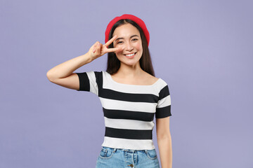 Smiling cheerful beautiful young brunette asian woman 20s wearing striped t-shirt red beret standing showing victory sign looking camera isolated on pastel violet colour background studio portrait.