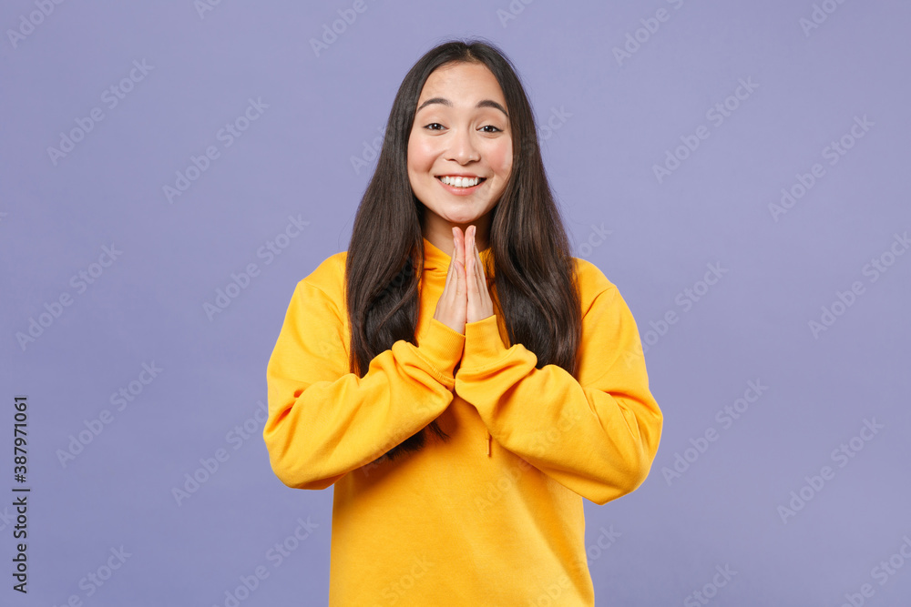 Wall mural Pleading smiling young brunette asian woman 20s wearing basic casual yellow hoodie standing holding hands folded in prayer looking camera isolated on pastel violet colour background studio portrait. - Wall murals