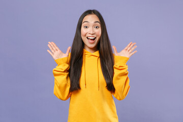 Surprised excited young brunette asian woman 20s in basic casual yellow hoodie standing keeping mouth open spreading hands looking camera isolated on pastel violet colour background studio portrait.