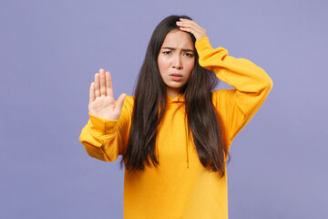 Dissatisfied displeased young brunette asian woman in casual basic yellow hoodie standing showing stop gesture with palm put hand on head isolated on pastel violet colour background studio portrait.