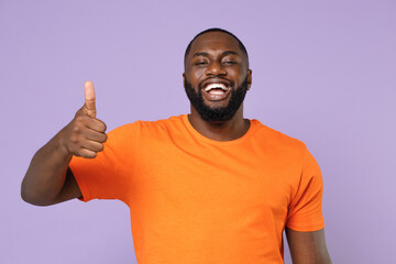 Cheerful funny young african american man 20s wearing basic casual orange blank empty t-shirt standing showing thumb up looking camera isolated on pastel violet colour background, studio portrait.