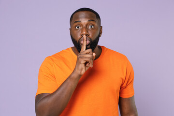 Secret young african american man 20s wearing basic casual orange blank empty t-shirt saying hush be quiet with finger on lips shhh gesture isolated on pastel violet colour background studio portrait.