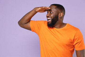 Fototapeta na wymiar Cheerful young african american man 20s wearing basic casual orange t-shirt standing holding hand at forehead looking far away distance isolated on pastel violet colour background, studio portrait.