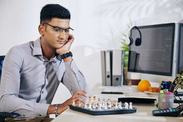 Pensive handsome young Indian software developer looking at chess board and thinking on next move