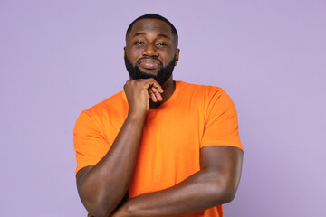 Confused puzzled young african american man 20s wearing basic casual empty orange t-shirt standing put hand prop up on chin looking camera isolated on pastel violet colour background, studio portrait.