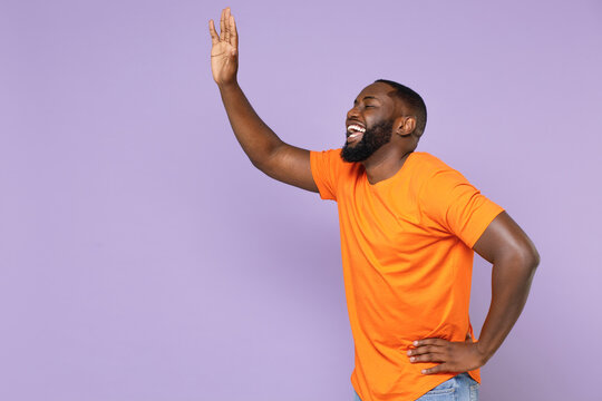 Side View Of Laughing Young African American Man In Basic Casual Orange T-shirt Standing Waving And Greeting With Hand As Notices Someone Isolated On Pastel Violet Colour Background, Studio Portrait.