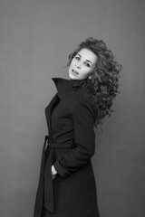 Beautiful slender brunette with long curly hair in a black coat in the studio on a gray background, bw