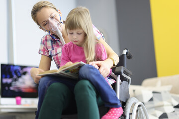 Woman in wheelchair and with an oxygen mask on her face is reading book with little girl. Helping...