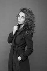 Beautiful slender brunette with long curly hair in a black coat in the studio on a gray background, bw