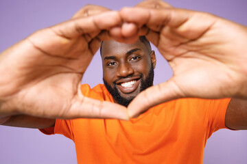 Smiling young african american man wearing basic casual orange blank empty t-shirt showing shape heart with hands, heart-shape sign looking camera isolated on pastel violet background studio portrait.