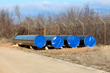 Four brand new large diameter pipes closed with nylon protection left at local oil pipeline construction site next to paved road and wire fence with barb wire on top surrounded with dense trees withou