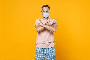Displeased young man in pajamas home wear face mask safe from coronavirus virus covid-19 showing stop gesture with crossed hands resting at home isolated on yellow background. Relax good mood concept.