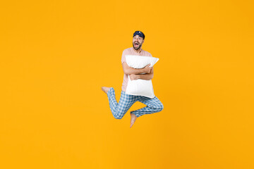 Full length of excited young man in pajamas home wear sleep mask jumping hold pillow keeping mouth open isolated on bright yellow colour background studio portrait. Relax good mood lifestyle concept.