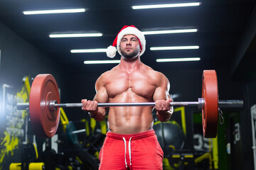 Fototapeta na wymiar Front view of a sexy muscular man wearing Christmas hat and red shorts lifting a barbell in a gym
