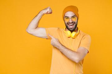 Strong funny young bearded man 20s wearing basic casual t-shirt headphones eyeglasses hat standing pointing index finger on biceps muscles isolated on bright yellow colour background, studio portrait. - obrazy, fototapety, plakaty