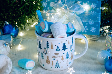 Obraz na płótnie Canvas Christmas cup with marshmallow on knitted white wool background and blue New Year decoration. Christmas garland lights on, cozy holiday atmosphere. 