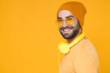 Side view of smiling cheerful funny young bearded man 20s wearing basic casual t-shirt headphones eyeglasses hat standing looking camera isolated on bright yellow colour background, studio portrait.