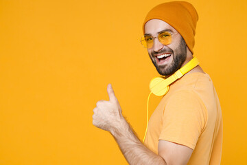 Fototapeta premium Side view of cheerful funny young bearded man 20s wearing basic casual t-shirt headphones eyeglasses hat standing showing thumb up looking camera isolated on bright yellow background, studio portrait.