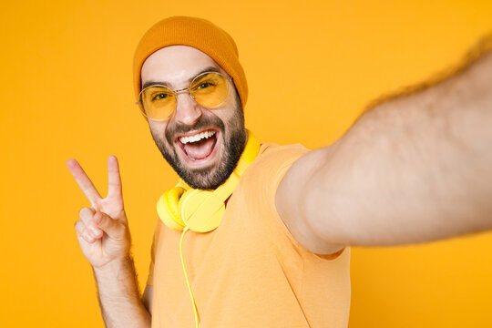 Close up of cheerful young bearded man in basic casual t-shirt headphones eyeglasses hat doing selfie shot on mobile phone showing victory sign isolated on bright yellow background studio portrait.