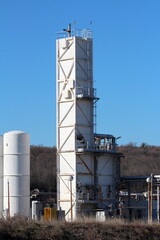 Tall old partially rusted white rectangle storage silo rising above row of brand new storage silos installed at the edge of modernized part of large oil refinery industrial complex surrounded with den