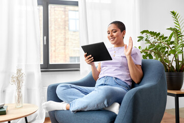 people, technology and leisure concept - happy young african american woman sitting in chair with tablet pc computer having video call and waving hand at home