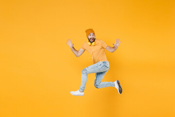 Fototapeta na wymiar Full length side view of excited shocked young man wearing basic casual t-shirt headphones hat jumping like running rising hands showing palms isolated on bright yellow background, studio portrait.