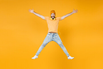 Fototapeta na wymiar Full length of excited surprised young man 20s wearing basic casual t-shirt headphones hat jumping spreading hands and legs looking camera isolated on bright yellow colour background, studio portrait.
