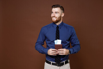 Smiling bearded young traveler tourist business man in blue shirt tie hold passport tickets looking aside isolated on brown background studio portrait. Achievement career wealth business concept.