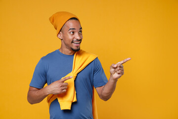 Smiling cheerful funny young african american man 20s in basic casual blue t-shirt hat pointing index fingers aside on mock up copy space isolated on bright yellow colour background, studio portrait.