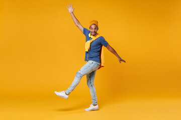 Full length side view of smiling cheerful young african american man 20s wearing blue t-shirt hat standing rising spreading hands and legs isolated on bright yellow colour background, studio portrait.