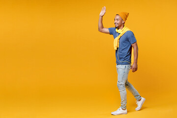 Fototapeta na wymiar Full length side view smiling joyful young african american man 20s in blue t-shirt hat waving and greeting with hand as notices someone isolated on bright yellow colour background, studio portrait.