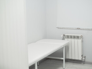 Fototapeta na wymiar White medical couch for examining and treating a patient in light clean doctor's cabinet. Window with curtain, battery in the room. Workplace