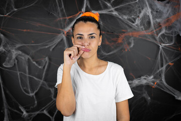 Young beautiful woman over black background with cobwebs and spiders mouth and lips shut as zip...
